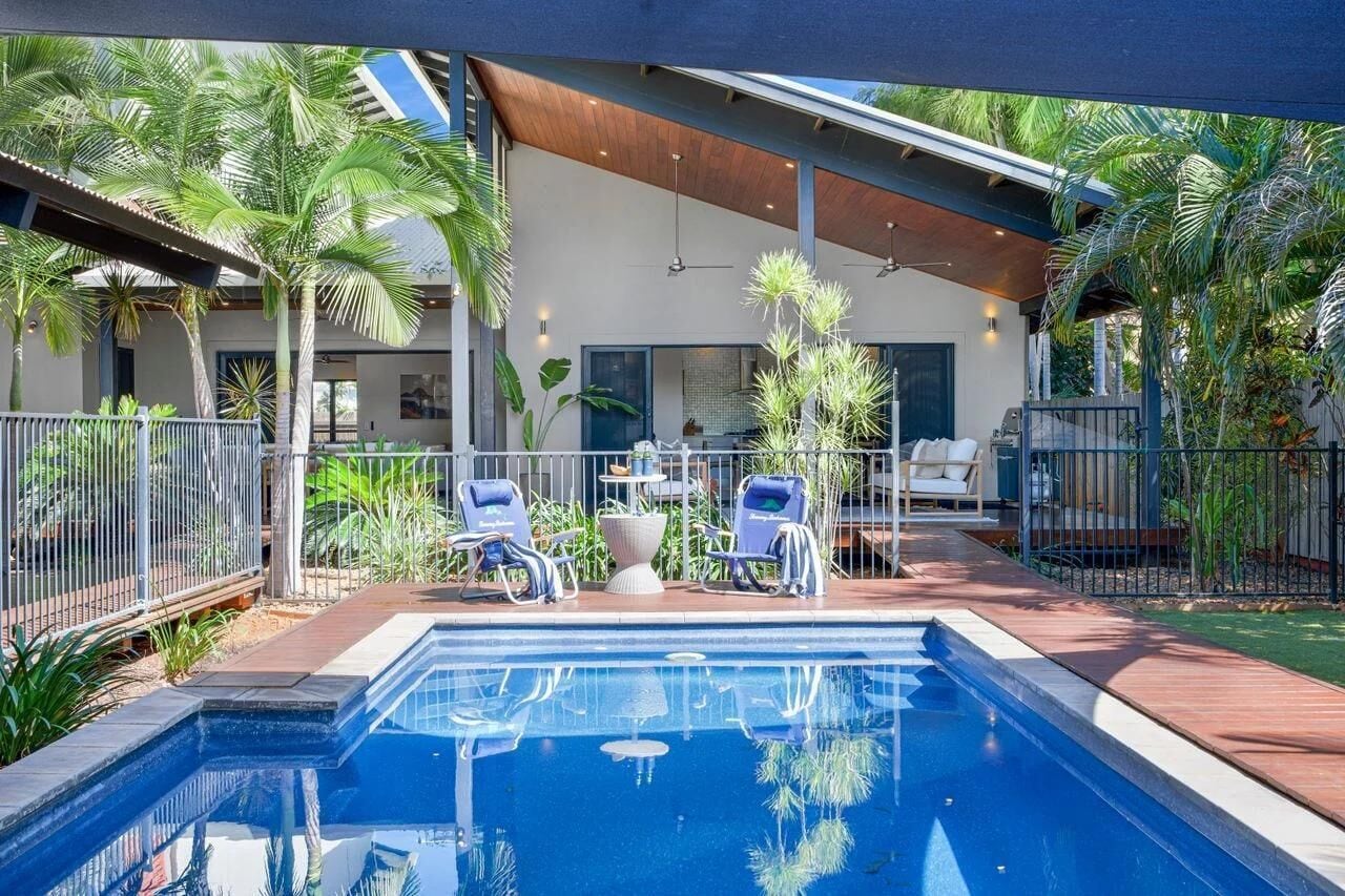 Private fenced pool area with blue loungers, tropical plants, and a covered patio with ceiling fans.