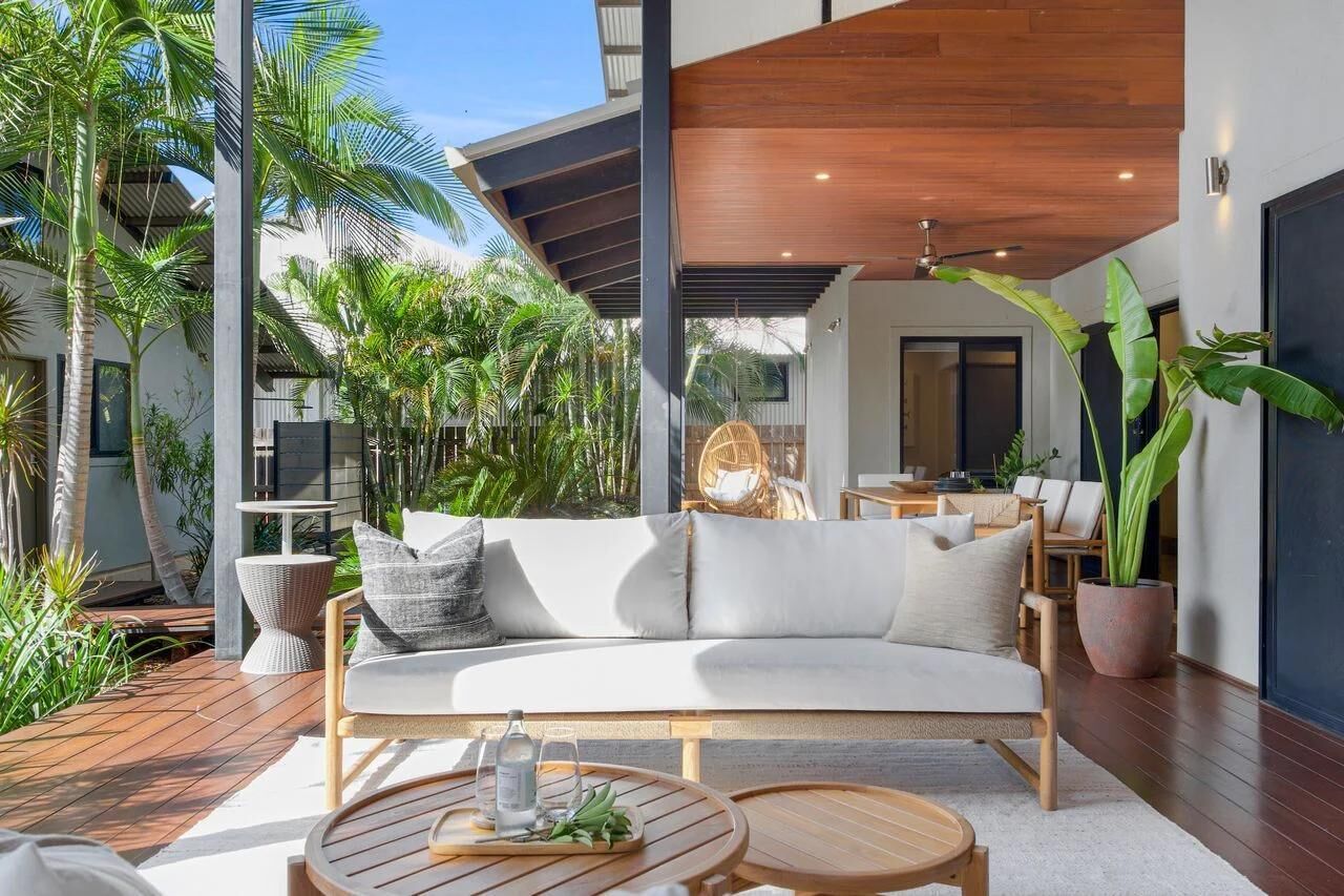 Outdoor lounge with light wood sofa, white cushions, round wooden tables, tropical plants, and covered patio ceiling.
