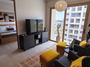A modern living room with a flat-screen TV, a sofa, ottomans, a bookshelf, and a large window with a view of buildings. - Apartment with city view in Zapopan, Jalisco (Zapopan)