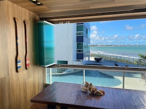 A modern room with a view of a pool and beach, wooden furniture, and a glass partition. - Bela Hospedagem - Imperial Bessa 121 (João Pessoa)