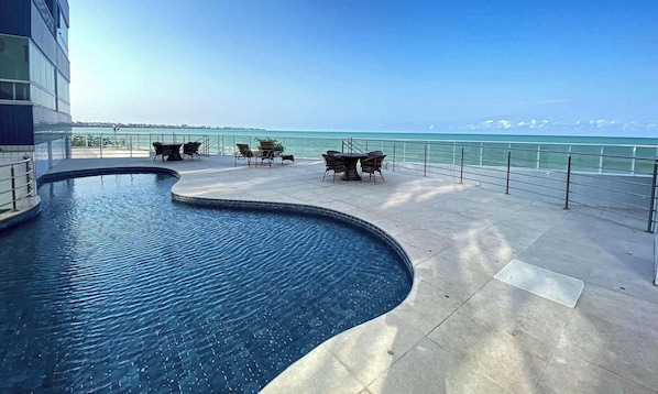 A pool with a curved edge, outdoor seating, and a view of the ocean. - Bela Hospedagem - Imperial Bessa 121 (João Pessoa)