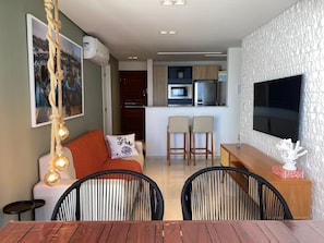 A modern living room with a sofa, two chairs, a dining table, a TV, and a kitchen in the background. - Bela Hospedagem - Imperial Bessa 121 (João Pessoa)