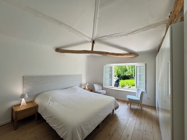 A bedroom with a large bed, a bedside table with a lamp, a window with a view of greenery, and a sloped ceiling with exposed beams. - In the quiet of the countryside - Authentic charm - Modern comfort - Air-conditioned (Touvérac)
