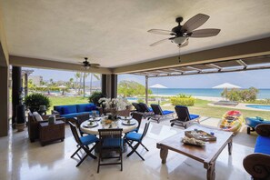 Ein offener Wohnbereich mit Esstisch, Stühlen und einer Couch. Deckenventilatoren, Meerblick und Sitzgelegenheiten mit Sonnenschirmen sind vorhanden.