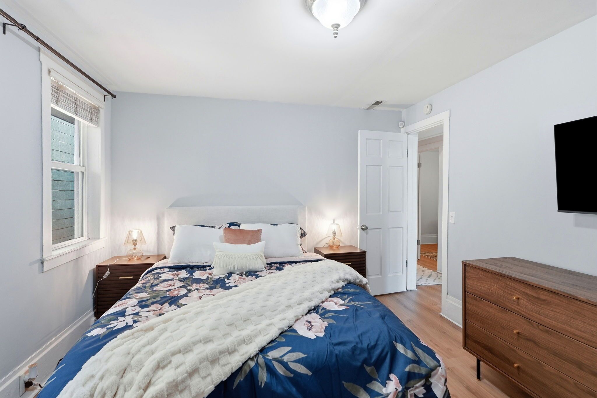 Un dormitorio con cama, mesitas de noche, cómoda, televisor y puerta que da a otra habitación.