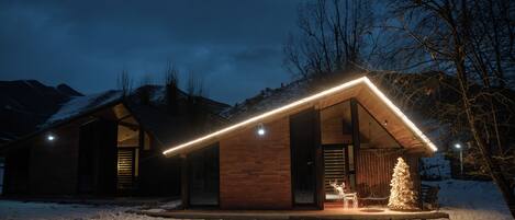 Une cabane en bois avec un toit en pente, un sapin de Noël illuminé et un banc dans la neige.