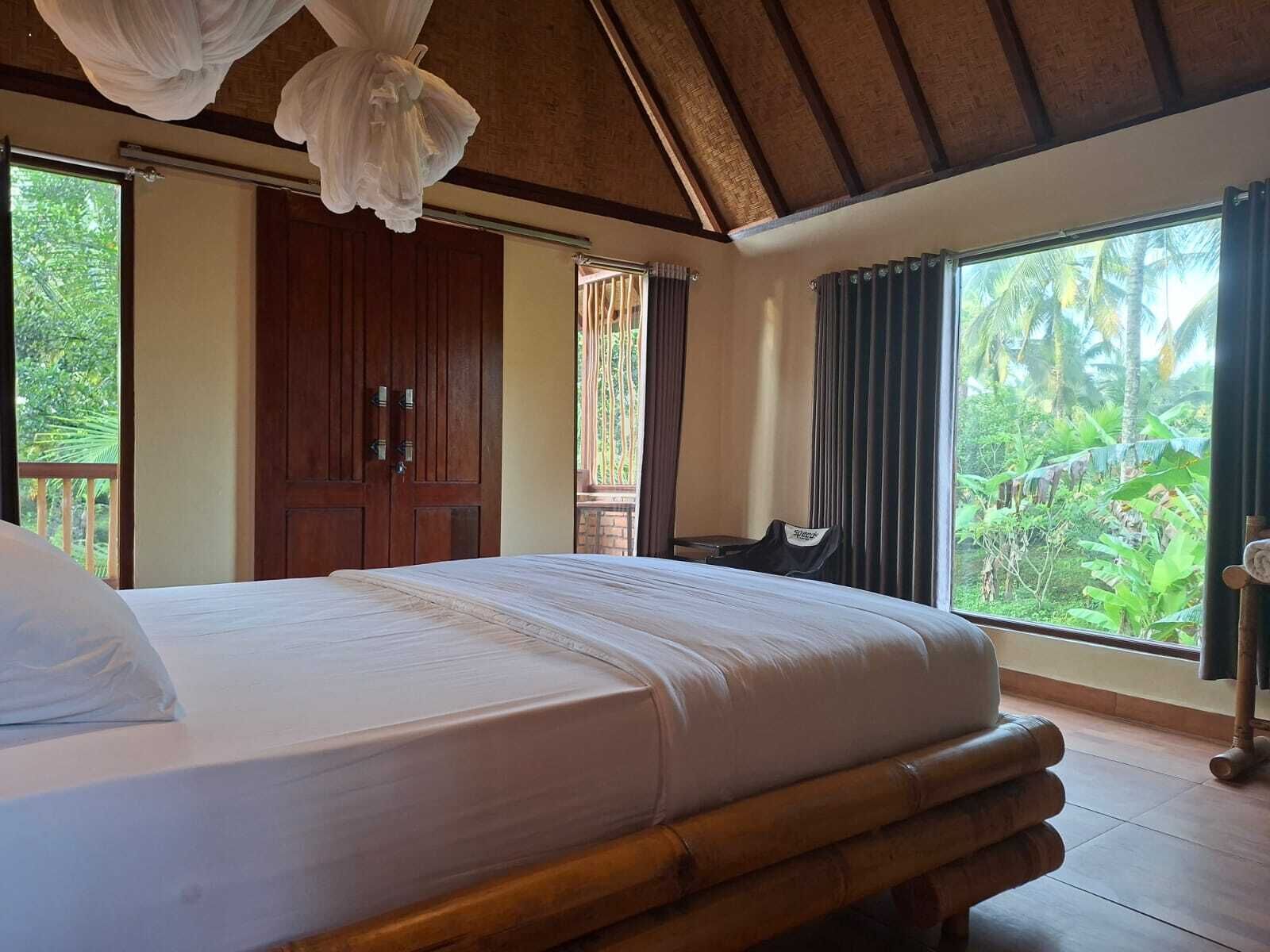 Quarto com uma cama grande, vigas de madeira no teto, vista para o verde e uma porta de madeira.