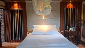 Una cama grande con sábanas blancas, una mesita de noche con una tetera y botellas, y un ventilador a la derecha.