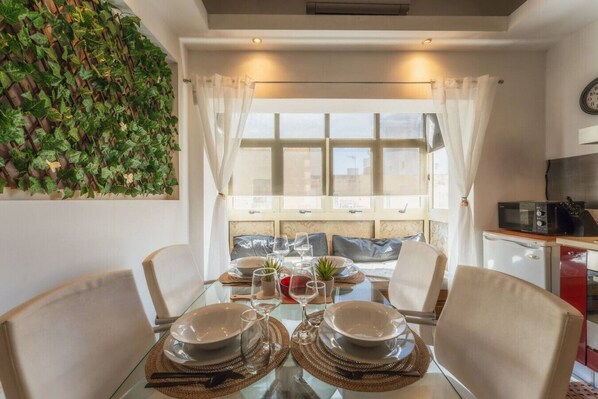 A dining area with a glass table set for four, a kitchen with a microwave, and a view through a window with sheer curtains. - Stylish Apartment | 1BR Near Sliema & Valletta (Gżira)