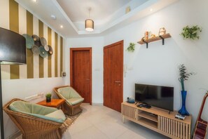 A living room with a TV, two wicker chairs, a coffee table, a wooden TV stand, and a striped wall decoration. - Stylish Apartment | 1BR Near Sliema & Valletta (Gżira)