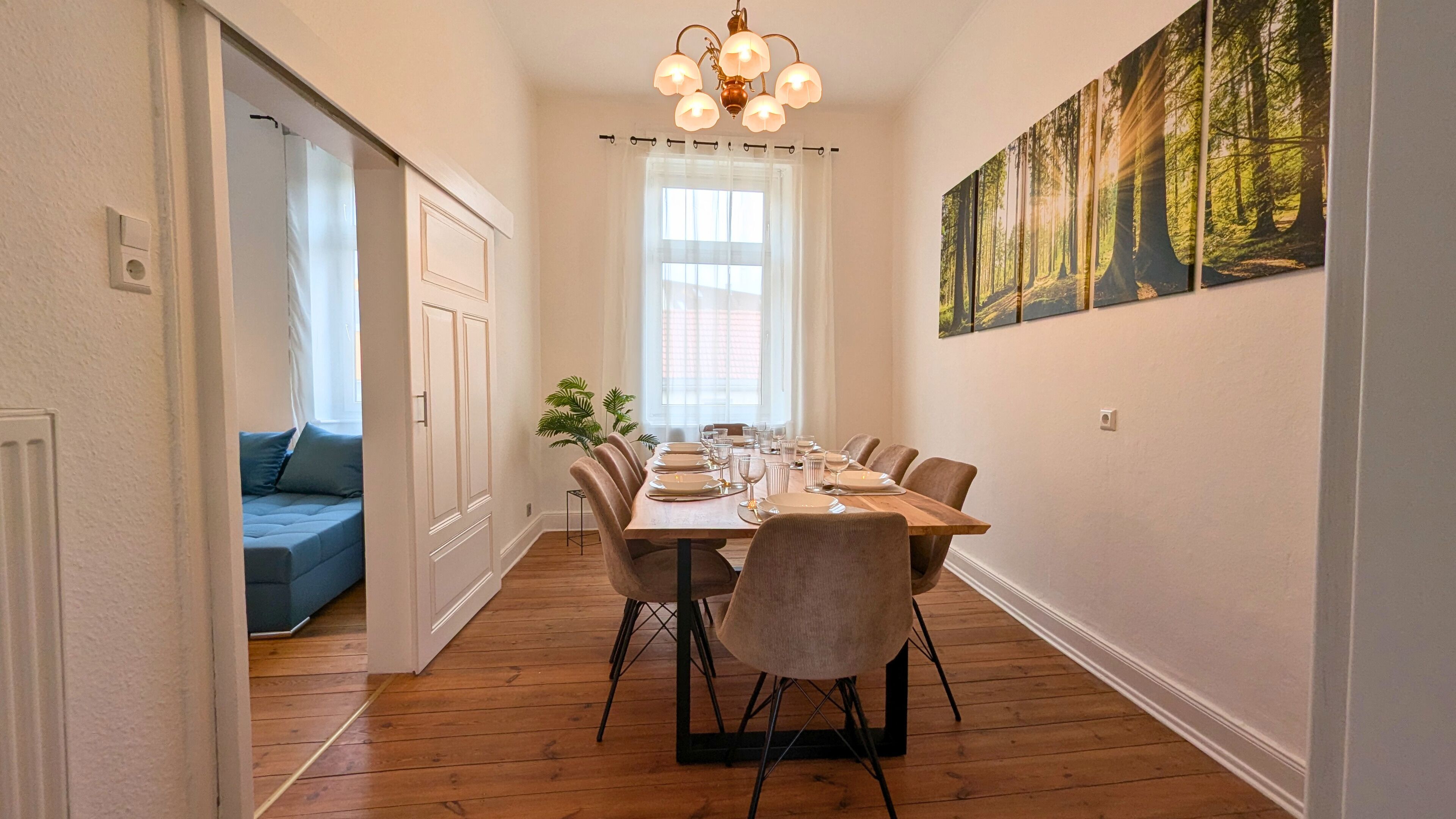 A dining area with a wooden table set for six, flanked by chairs with black metal legs. A large window with sheer curtains allows natural light, and a forest-themed painting adorns the wall.