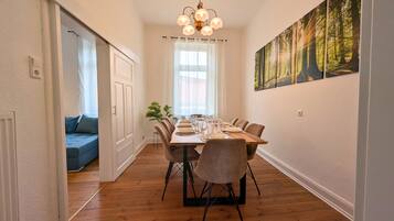 A dining area with a wooden table set for six, flanked by chairs with black metal legs. A large window with sheer curtains allows natural light, and a forest-themed painting adorns the wall.