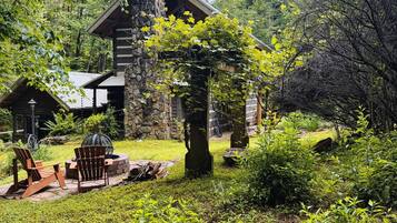 A rustic cabin with a stone chimney, surrounded by lush greenery and a well-maintained garden with Adirondack chairs.