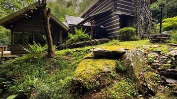 A rustic cabin with a stone chimney, surrounded by lush greenery and a forested backdrop.