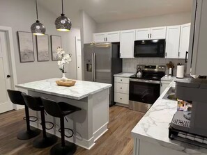 A modern kitchen with white cabinets, a marble island, black bar stools, stainless steel appliances, and pendant lighting.