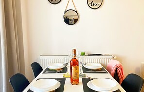 Une table à manger est dressée avec des assiettes, une bouteille de vin et des verres. Des chaises sont disposées autour de la table, et on aperçoit un radiateur à l’arrière-plan.