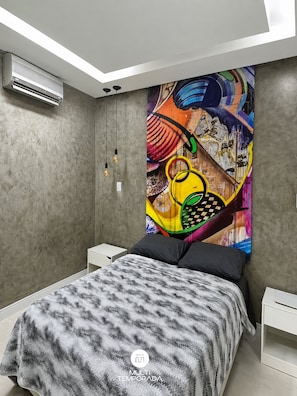 A modern bedroom with a bed, a colorful abstract painting, a ceiling light, and a wall-mounted air conditioner.