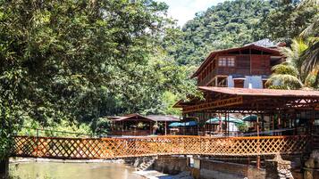 A wooden building with a balcony, surrounded by lush greenery and a bridge over a body of water.