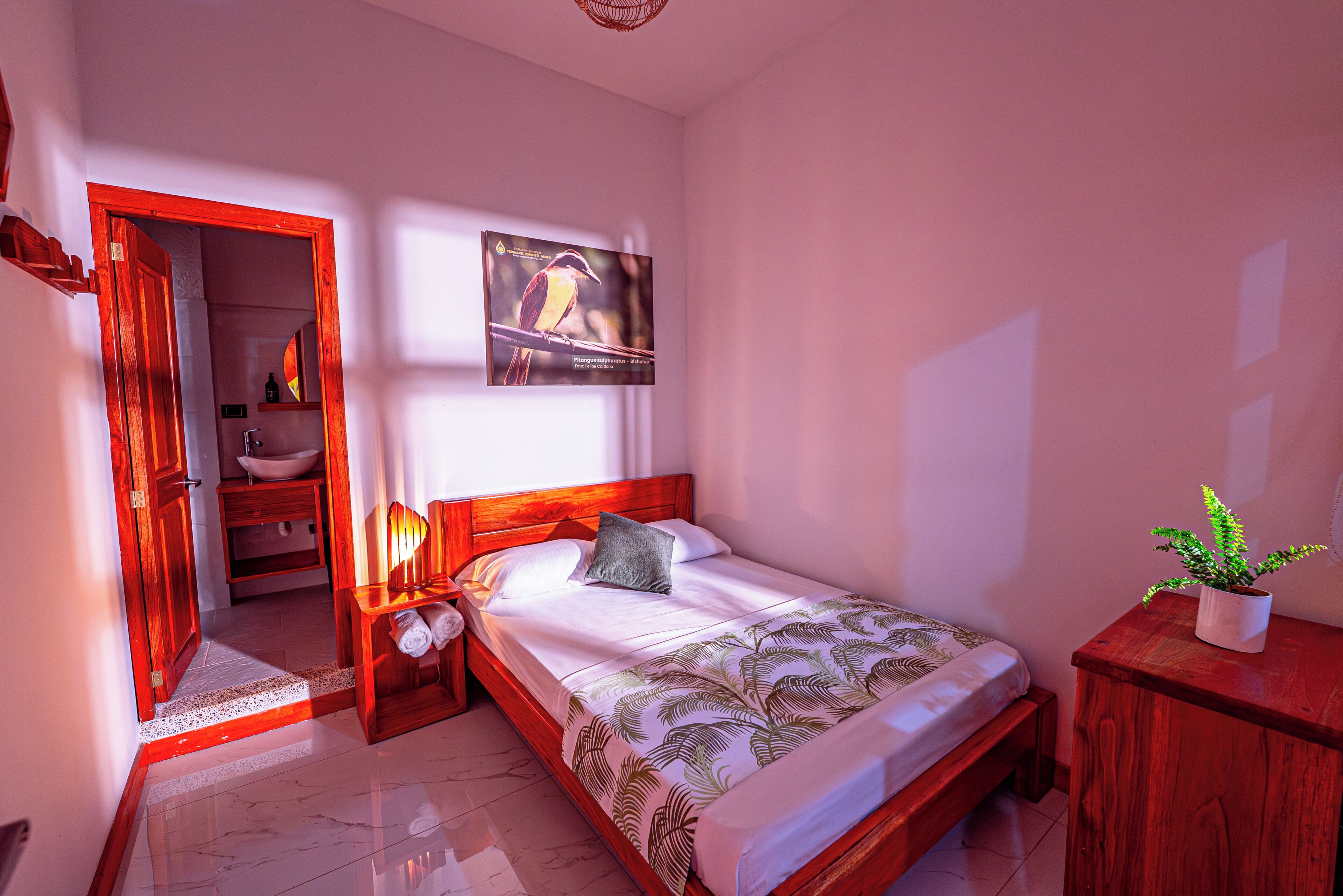 Quarto com cama, mesa de cabeceira, cabeceira de madeira, quadro na parede, planta em vaso e banheiro visível por uma porta aberta.