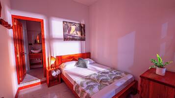 Quarto com cama, mesa de cabeceira, cabeceira de madeira, quadro na parede, planta em vaso e banheiro visível por uma porta aberta.