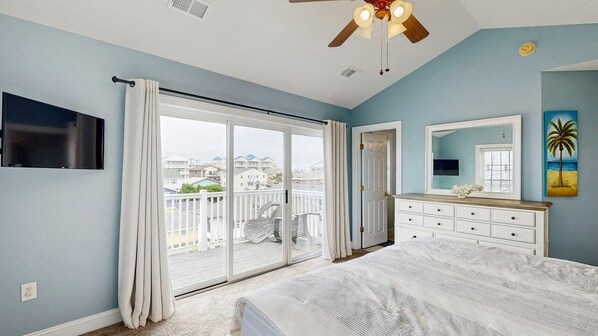 A bedroom with a large bed, a ceiling fan, a TV, a dresser, and a sliding glass door leading to a balcony. - Spacious coastal townhouse with multiple decks, gas fireplace, & relaxing Bay views (Ocean City)