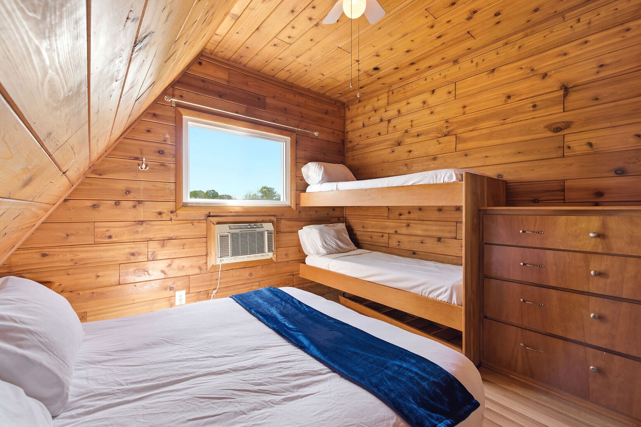 A room with wooden walls, a bunk bed, a window, a dresser, and an air conditioning unit.