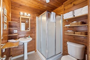 Baño de cabaña de madera con ducha, lavamanos, inodoro y estantes con toallas.
