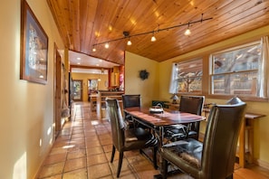 A dining area with a wooden table and chairs, a kitchen in the background, and a window with curtains. - Bluebird Day - Desert Zen in Bend with 2 queens, fenced yard, fireplace, Walk Everywhere! (Bend)