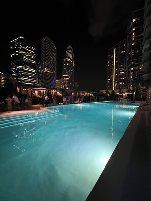 Une piscine avec des marches permettant d’y accéder, entourée d’immeubles dont les fenêtres sont illuminées.