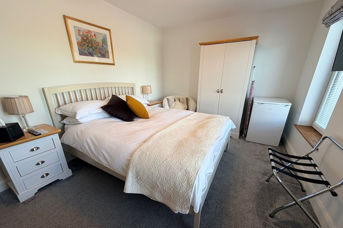 Ein Schlafzimmer mit Bett, Nachttisch, Lampe, Kleiderschrank und einem kleinen Kühlschrank.