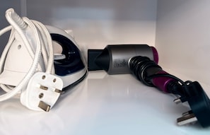 A white iron, a black and pink hairdryer, and a white power adapter on a white surface. - Ardbrin Lodge Guest Accommodation  (Antrim)
