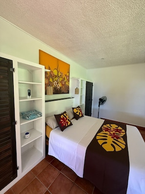 Habitación de hotel con cama, armario, estantería, un cuadro y ventilador.
