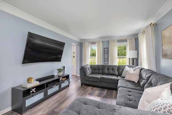 Ein Wohnzimmer mit einem Ecksofa, einem Flachbildfernseher, einer dunkelholzartigen Unterhaltungsanlage und einem großen Fenster mit Vorhängen.