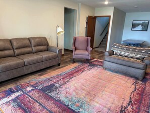 Un salon avec un canapé en cuir marron, un fauteuil à motifs, un tapis coloré, une lampe sur pied et un escalier en arrière-plan.