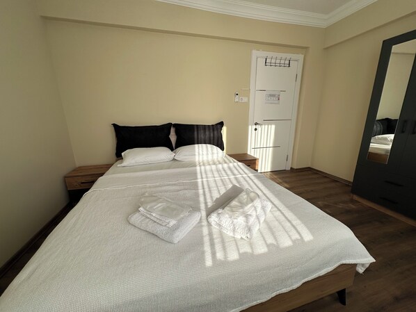 Una cama bien hecha con una colcha blanca y dos almohadas, una mesita de noche de madera con una pequeña mesa, una puerta blanca con manija y un espejo en la pared.