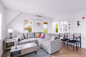 Ein modernes Wohnzimmer mit einem Ecksofa, einem Couchtisch, einem Schreibtisch mit Stühlen und einem Fenster mit Blick auf Bäume.