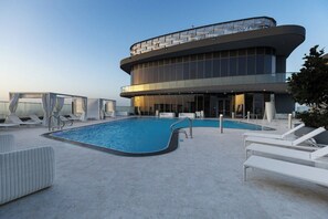 Ein Pool auf dem Dach mit Liegestühlen, ein gläsernes Gebäude und ein klarer Himmel.