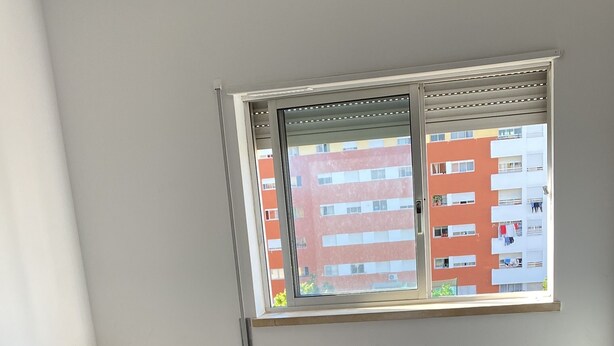 Ein Schlafzimmer mit Bett, einem Fenster mit Jalousien und Blick auf ein Gebäude.