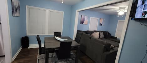 A living room with a dining table, chairs, a sofa, a ceiling fan, and a television mounted on the wall.