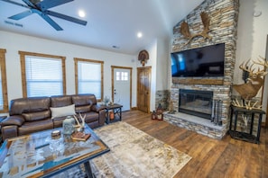 A living room with a stone fireplace, leather sofa, and a ceiling fan. - Luxe Country Lodge | Fire Pit & Acreage (McLouth)