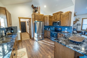 A kitchen with wooden cabinets, stainless steel appliances, a marble countertop, and a rustic decor. - Luxe Country Lodge | Fire Pit & Acreage (McLouth)