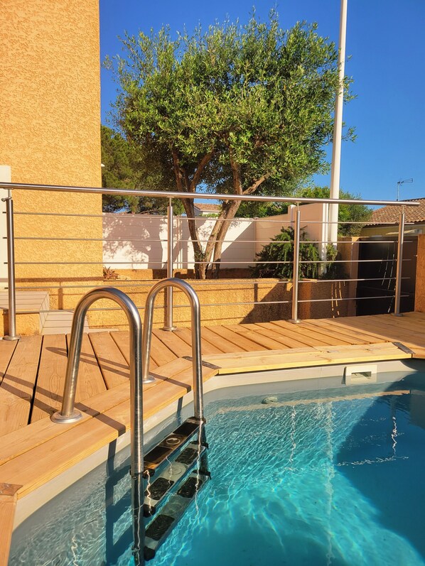 A swimming pool with a wooden deck and metal railings, surrounded by a building with a textured wall and a tree. - 🌿 Villa L'Olivier Private pool, garden, 8 people, air conditioning and beaches 4 km away 🌊 (Portiragnes)