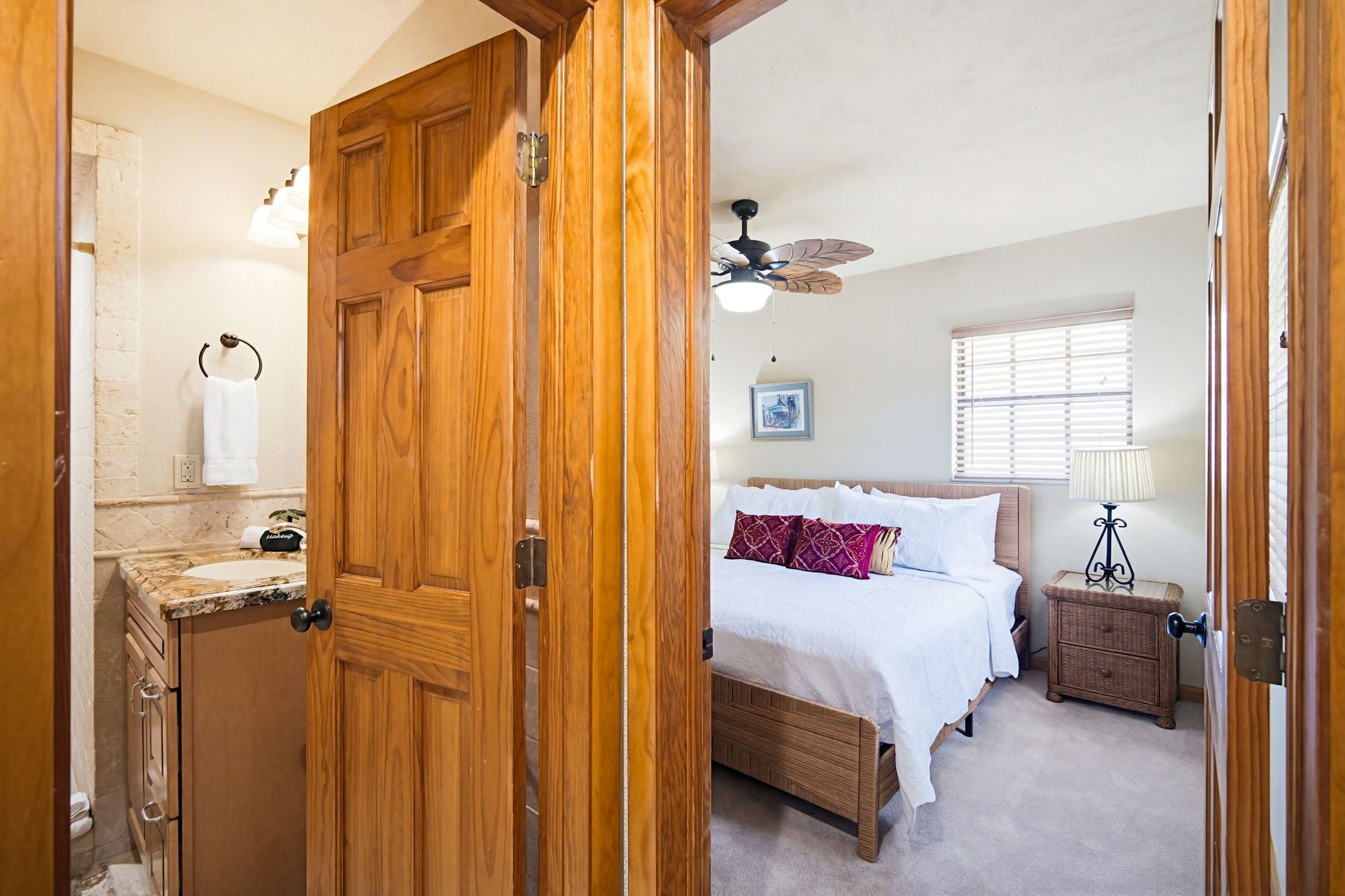 Une chambre à coucher comprenant un lit, une table de chevet, une lampe, un ventilateur de plafond, une fenêtre avec des stores et une salle de bain avec un lavabo et un porte-serviettes.