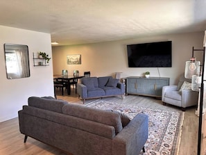 Ein Wohnzimmer mit einer Couch, einem Sessel, einem Fernseher und einem Essbereich mit Stühlen und einem Tisch.