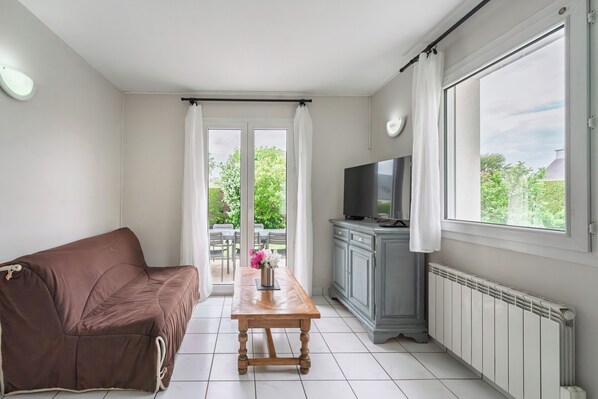 A living room with a sofa, a wooden table with a vase, a TV, and a cabinet. - L'Azalée - Petfriendly - Terrasse (Saint-Méloir-des-Ondes)