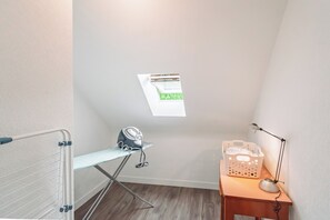 Une chambre d’angle avec un bureau en bois, un panier à linge, une lampe de bureau et une fenêtre donnant sur un espace extérieur verdoyant.