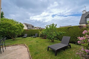Un jardin avec une pelouse, deux chaises longues, un petit espace aménagé en jardin et une haie.