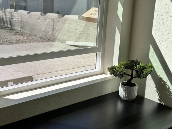 Een raam met uitzicht op een bouwplaats, een klein bonsaiboompje in een pot op de vensterbank, en een structuurrijke muur.