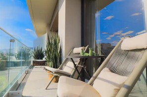 Een balkon met glazen hekwerk, een lounge stoel, een klein tafeltje en potplanten.