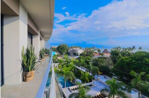 A balcony with a pool, potted plants, and a view of a coastal area with buildings and palm trees. - Luxury 2BR w/ Ocean Views • Gym • Jacuzzi • Infinity Pool • Walk Everywhere (Bucerías)